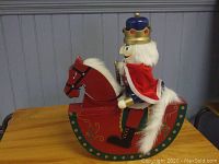 Front angled view of the wooden nutcracker wearing a blue crown and red cape on a red rocking horse with decorative painting and white fur mane/tail.