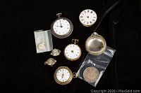 All items arranged on black background showing one Canadian silver dollar coin in plastic bag, five pocket watches of different sizes and styles, a loose watch face, and a silver case.