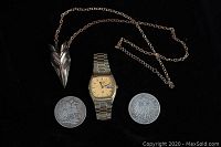 Front view of Seiko watch, copper raven pendant necklace, 1889 silver crown coin, and 1903 German mark coin on black background