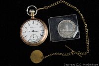 Elgin gold-tone pocket watch, white dial, black numbers, linked chain with brass fob, and 1939 Canadian silver dollar coin in plastic holder all shown on black background.