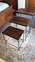 Three rectangular nesting tables with wooden plank tops and black metal legs arranged stacked and side by side on a carpeted floor in front of a wooden bed frame.