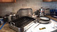Wide view of roasting pan with wire rack, stock pot, and assortment of fry pans and pots in kitchen setting