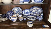 Various Willow pattern china items displayed in a cabinet including plates, pitchers, bowls, salt and pepper shakers.