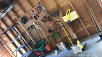Scotts lawn spreader next to a shovel and black shelving, wooden garden rake mounted above, various garden tools hanging on pegboard