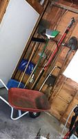 Red wheelbarrow in garage leaning on the wall with wooden handled garden tools visible behind