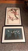 Both framed art pieces, cherry blossoms on top and flying cranes below, shown on carpeted floor.