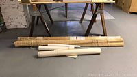 Photo of two bamboo outdoor screens and two wooden sawhorses with some wallpaper rolls on floor near a table.