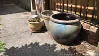 Four outdoor pots on concrete ground next to wooden fence. Includes large blue glazed terracotta pot, beige plastic pots, small painted floral pot inside one beige pot.