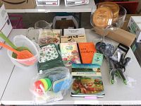 Overview of entire lot showing wooden bowls stacked in a glass bowl, various cookbooks, unopened kitchen linens package, spatulas, scrubbers, and glass pitcher on a white surface.