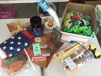 Photo of boxed mixed decor including folded American flag, blue glass vase, wooden fall cutouts, candles, ribbons, and other decorative items on table.