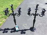 Pair of black metal candelabra standing upright outdoors on paved driveway with green grass and bushes in the background. Both candelabra have 5 candle holders each and stand 31 inches tall.