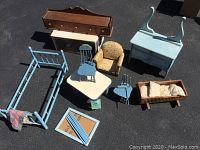 Overview photo showing seven pieces of doll furniture on pavement including bed, cradle, table with chairs, dresser, club chair, shadow box, and storage shelf.