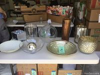 Nine pieces of decorative glass arranged on a white table including various vases, bowls, and a plate with reflective and textured finishes.