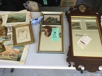 Overview of the lot items on the table including antique mirror with reverse painted top, multiple small prints, and ceramic animal creamers