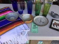 Photo showing colorful glassware including tall green, blue, purple glasses, several pottery bowls, a carved bird sculpture, a framed art piece, and a folded colorful woven textile runner.