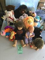 Full front group listing showing American Girl doll in navy dress in center with baby doll, Tigger Beanie Baby, Ganz white elephant, lion, hedgehog, gorilla, shark plush and other stuffed animals arranged behind