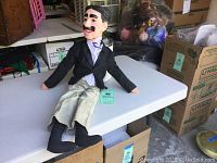 Full body photo of Groucho Marx ventriloquist mannequin sitting on a table in a cluttered room.
