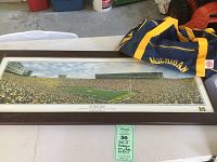 Framed panoramic scenic print of Michigan Stadium with blue and maize Michigan duffle bag beside it