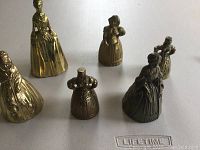 Six brass and brass-colored metal lady figurine bells standing on a white surface, showing size and detail up close