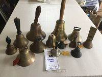 Overview of thirteen classic handbells varying in size and handle style, displayed on a tabletop.