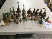 Full view of all 23 vintage bells arranged on a white plastic table, showing variety of sizes and shapes from small to large, some with tags attached.