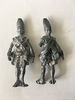 Front view of two vintage metal soldier figures standing upright on a white surface, showing detailed uniforms and tall hats.
