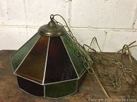 Photo showing hexagonal stained glass lamp with green and burgundy textured glass panels, metal top, hanging chain, and electrical cord.