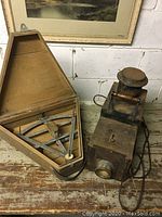 Wide shot of both vintage items on a wooden surface against a white brick wall, including the surveyor tool in its case and the oil lantern.