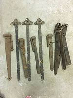 Photo showing 6 large vintage metal hinges arranged on a flat surface with visible wear, paint chipping, and rust spots.