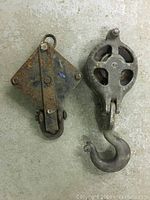 Photo showing two large vintage metal pulleys placed side by side on a concrete floor. One pulley has a large hook underneath, the other a more rectangular body with a swivel hook.