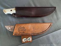 Pair of hunting knives sheathed in brown and tan leather with decorative elements, laid flat on grey fabric.