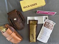 Top view showing Buck Knife original yellow box, Buck Knife use & care pamphlet, linked pamphlets, and three sheaths: two brown woven leather and one tan leather with stitching.
