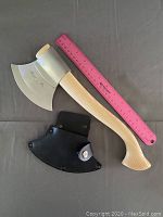 Full view of the throwing axe with wooden handle and stainless steel blade next to a pink ruler for scale, also showing the black leather sheath.