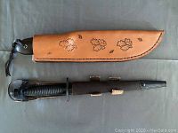 Two hunting knives displayed side by side, one in tan leather sheath with embossed leaves, the other in black leather sheath with ribbed handle.