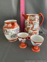 Photo showing the teapot, small pitcher, and two cups with a pink ruler for size reference. The items feature orange and cream coloring with floral and bird patterns.