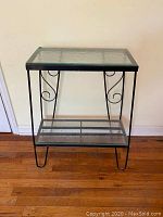 Front view of black iron plant stand with frosted glass top and bottom shelves on hardwood floor.