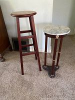 Two tall wooden plant stands side by side on carpet. One has a white marble top; the other has a painted animal wooden top.
