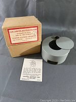 Cylindrical gray metal carrying case with hinged lid, original box and instruction leaflet