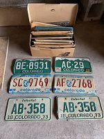 Box filled with approximately 36 vintage Colorado license plates, with 6 plates displayed outside the box showing various years and designs.