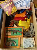 Box showing a variety of vintage toy guns including colorful plastic pistols and boxed metal toy gun replicas