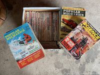 Overview photo showing an open box filled with vintage Popular Mechanics magazines and several loose magazines laid out around it on a concrete floor.