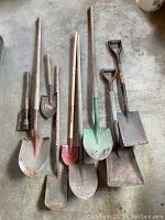 Nine old garden shovels and tools laid on concrete floor, showing variety of blade shapes and handle lengths.