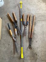 Photo of four pruning shears with wooden and plastic handles and a heavy duty crowbar with yellow ends, arranged on concrete floor.
