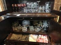 Wide view of wooden cabinet showing various clear glassware on top shelves, amber glassware and cut glass dishes inside the cabinet, and drawer of linens open below.