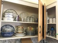 Kitchen cabinet shelf with stacked dinner plates, bowls, and glassware including mugs and glasses.