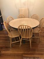 Round solid wood dining table with six matching chairs around it and a leaf placed behind against the wall.
