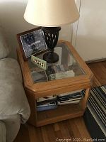Side view of solid oak side table with glass top and bronze color decorative table lamp with beige shade, positioned next to a sofa and adjacent to a wall.