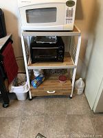 Photo of metal utility cart with drawer and two shelves, holding a white Sharp microwave on top and a black toaster oven on middle shelf, kitchen setting