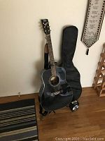 Full view of black Yamaha F335 acoustic guitar standing on a black guitar stand with soft gig bag, guitar strap, and clip-on tuner placed nearby.