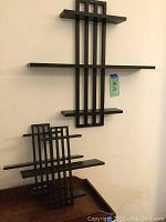 Photo showing larger wall shelf mounted on wall and smaller shelf leaning against wall, depicting geometric vertical slats and rectangular horizontal shelves in black paint.